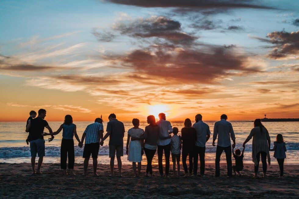 La imagen muestra a una familia numerosa. Están cogidos de la mano y de pie junto al mar. La familia contempla una puesta de sol.