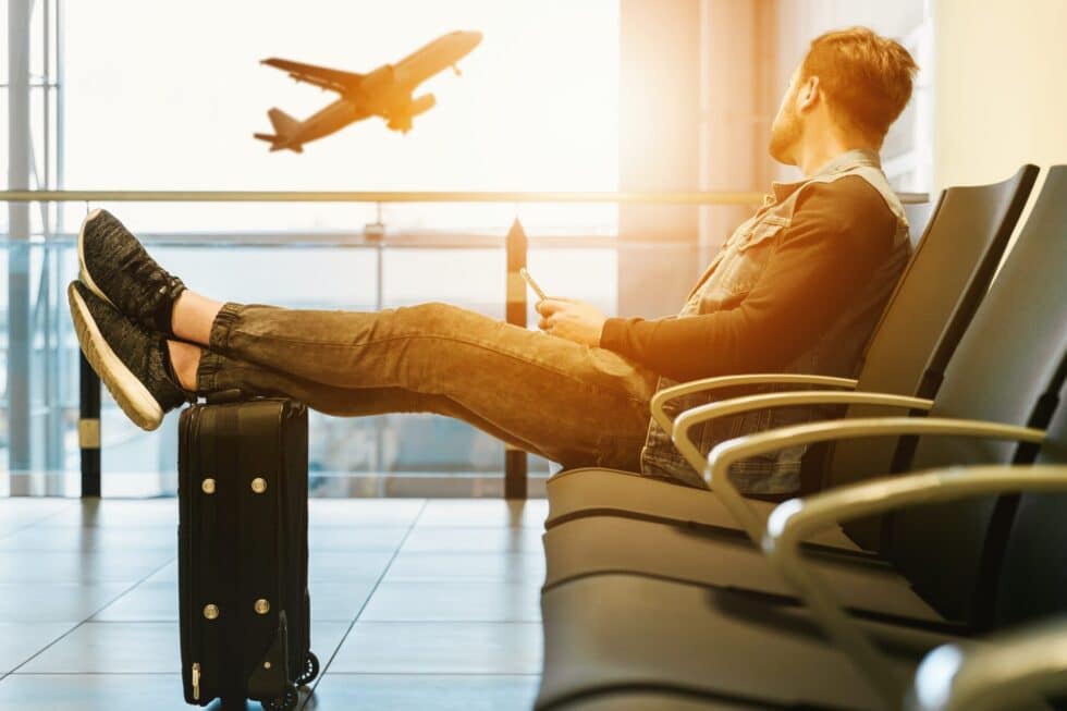 A young man sits in an airport hall before traveling to his home country. His feet are resting on the suitcase. He looks out of the window and sees an airplane taking off.