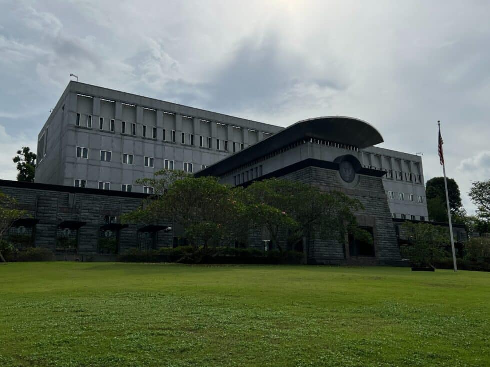 A imagem mostra um edifício da embaixada cinzento com uma bandeira dos EUA em primeiro plano. À sua frente, um prado verde com árvores verdes.