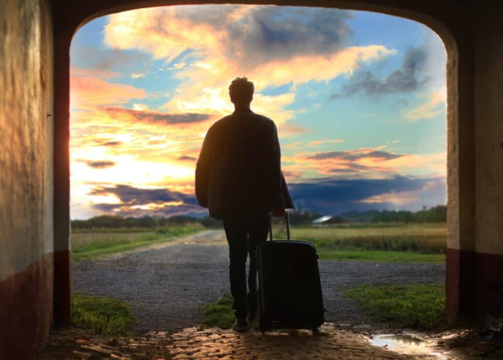 Sur la photo, on voit un homme. Il se tient avec une valise à la sortie d'un portail. Devant lui se trouve un paysage de prairie avec un ciel bleu.