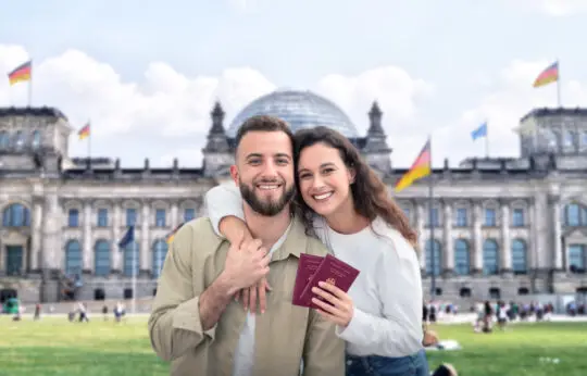 Ein glückliches Paar steht vor dem Reichstagsgebäude in Berlin und hält stolz ihre deutschen Pässe in den Händen. Das Bild symbolisiert den erfolgreichen Abschluss des Einbürgerungsprozesses und verdeutlicht die Erfüllung der Einbürgerungsvoraussetzungen in Deutschland.