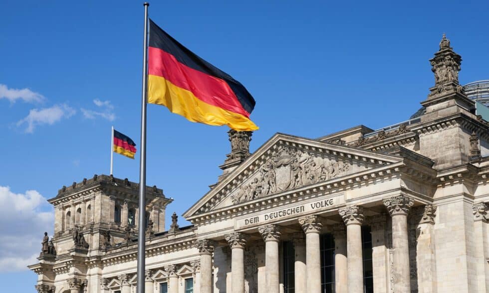 Zdjęcie przedstawia niemiecką flagę przed budynkiem Reichstagu w Berlinie. Niebo w tle jest niebieskie