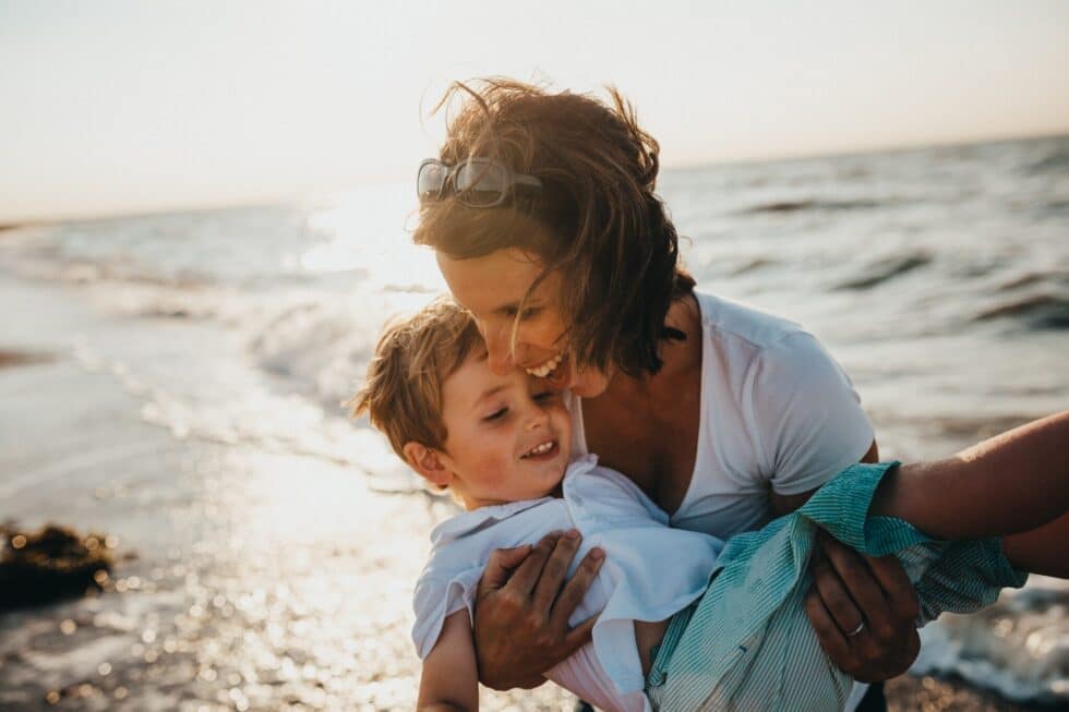 Auf dem Bild ist eine Mutter mit ihrem Sohn zu sehen. Sie befinden sich an einem Strand. Im Hintergrund sind die Wellen des Meeres zu sehen