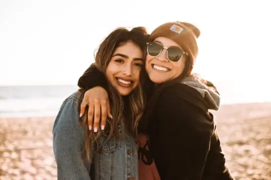 Auf diesem Bild umarmen sich zwei Freunde. Im Hintergrund ist ein Strand und das Meer zu sehen. Eine Verpflichtungserklärung für Besucher bietet dem Einlader Chancen aber auch Risiken.