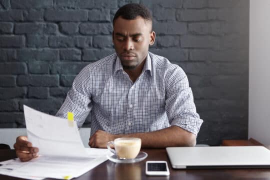 一名认真的非裔美国男子在咖啡馆喝咖啡休息时研究财务文件。他坐在办公桌前，拿着一台笔记本电脑、一部智能手机和一杯咖啡，一边翻阅文件，可能是为了申请入籍。