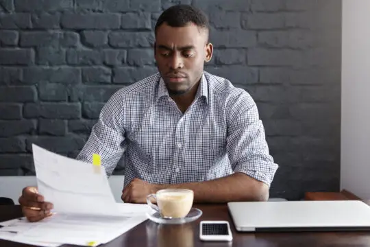 Ein ernsthafter afrikanisch-amerikanischer Mann studiert finanzielle Unterlagen bei einer Kaffeepause in einem Café. Er sitzt an einem Schreibtisch mit einem Laptop, einem Smartphone und einer Tasse Kaffee, während er Dokumente durchgeht, möglicherweise für seinen Einbürgerungsantrag.