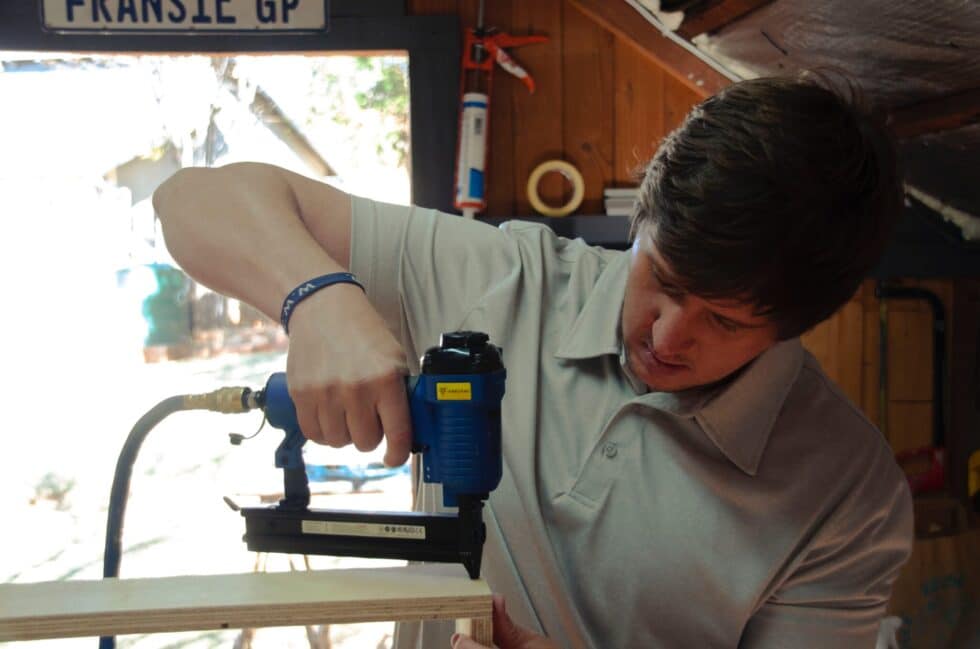 In questa immagine si vede un apprendista artigiano al lavoro. Ha un trapano in mano e sta forando un pezzo di legno.