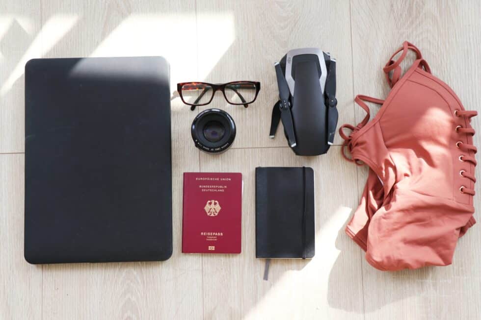 Auf diesem Bild ist ein deutscher Pass mit Reisesachen zu sehen. Zu den Reisesachen gehören ein Damenoberteil, eine Brille, ein Notizbuch, eine Kamera und ein Tablet.