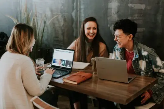 Auf diesem Foto sieht man junge Leute am Tisch, die miteinander lachen. Es sind zwei Frauen und ein Mann. Die Einbürgerung als Student ist unter bestimmten Voraussetzungen möglich.