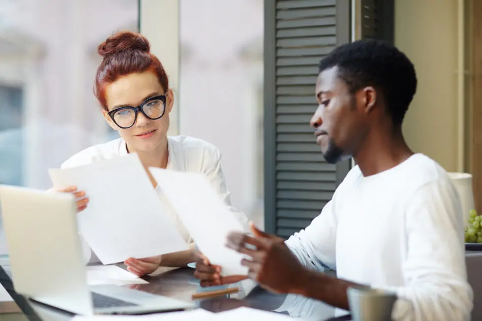 Ein Mann und eine Frau sitzen in einem Büro und besprechen gemeinsam Dokumente. Sie wirken konzentriert und engagiert, während sie wichtige Unterlagen durchgehen, die möglicherweise im Zusammenhang mit einem Einbürgerungsantrag stehen. Ein Laptop steht auf dem Tisch und unterstützt die professionelle Arbeitsumgebung.