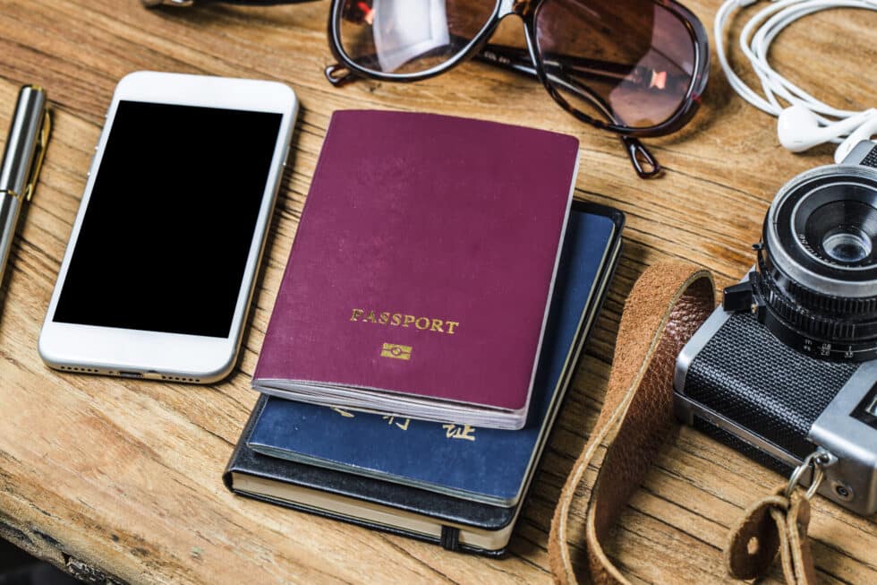 Auf diesem Foto sehen Sie unterschiedliches Reisezubehör. Ein deutscher Pass, ein Handy, eine Fotokamera und eine Sonnenbrille. Alles liegt auf einem Tisch