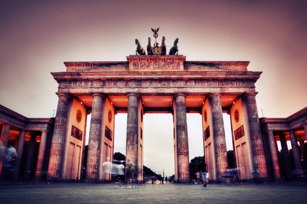Auf diesem Foto sehen Sie das Brandenburger Tor in Berlin im Sonnenlicht. Vor dem Tor befinden sich ein paar Touristen die Fotos machen
