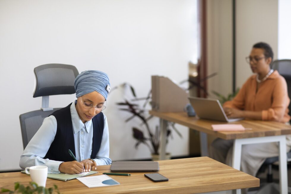 Auf diesem Foto arbeiten zwei Frauen an jeweils einem Schreibtisch. Eine der Frauen trägt Kopftuch und notiert etwas auf einem Blatt Papier. Die andere Frau tippt etwas in ihren Laptop ein.