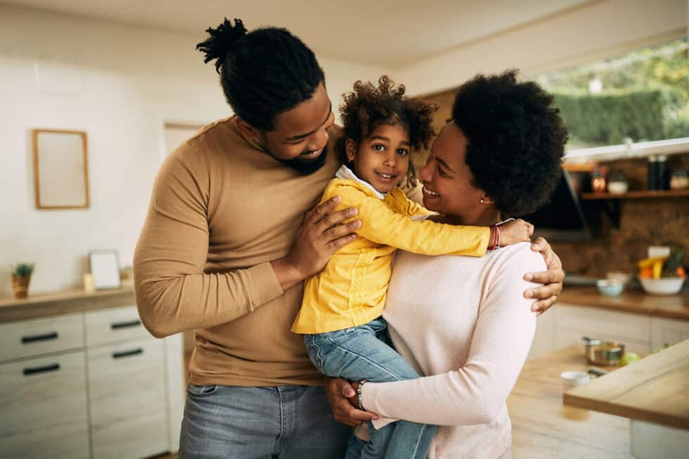 Auf diesem Bild ist eine fröhliche afroamerikanische Familie in Ihrer Wohnung zu sehen. Im Hintergrund befindet sich eine Küche.