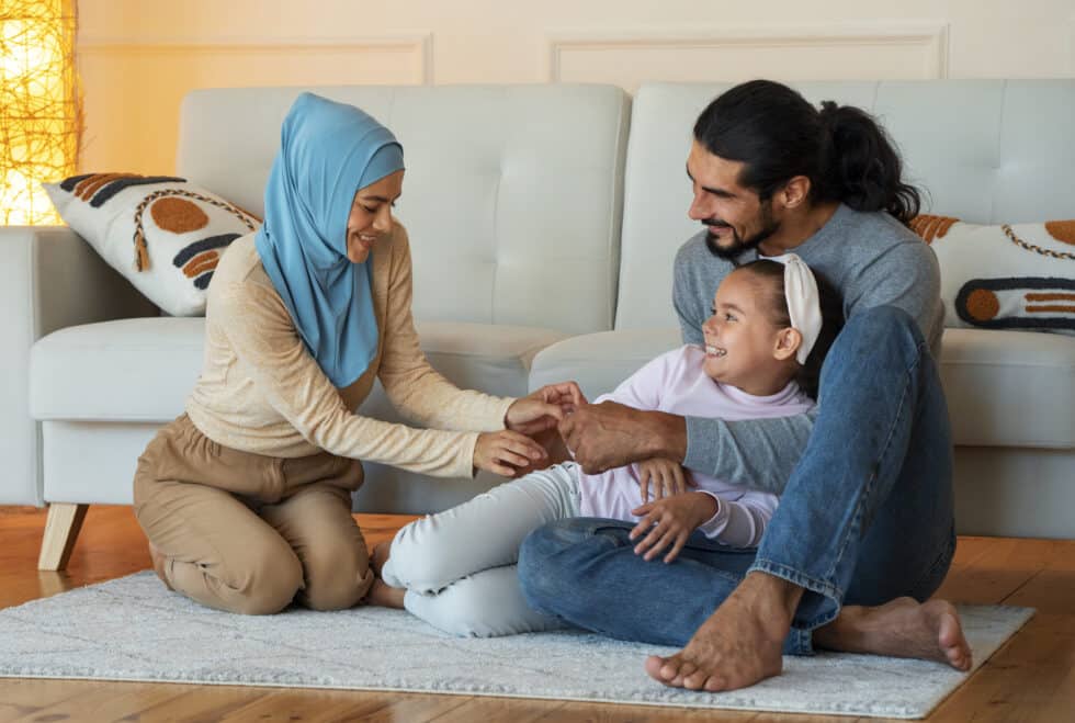 Auf diesem Foto sehen Sie eine junge muslimische Familie, die auf dem Boden ihrer Wohnung sitzt. Im Hintergrund befindet sich eine Couch