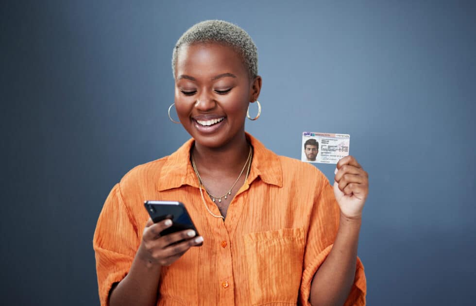 Auf diesem Foto sehen Sie eine lächelnde Frau, die in einer Hand einen Aufenthaltstitel 25b AufenthG trägt und in der anderen ein Mobiltelefon.