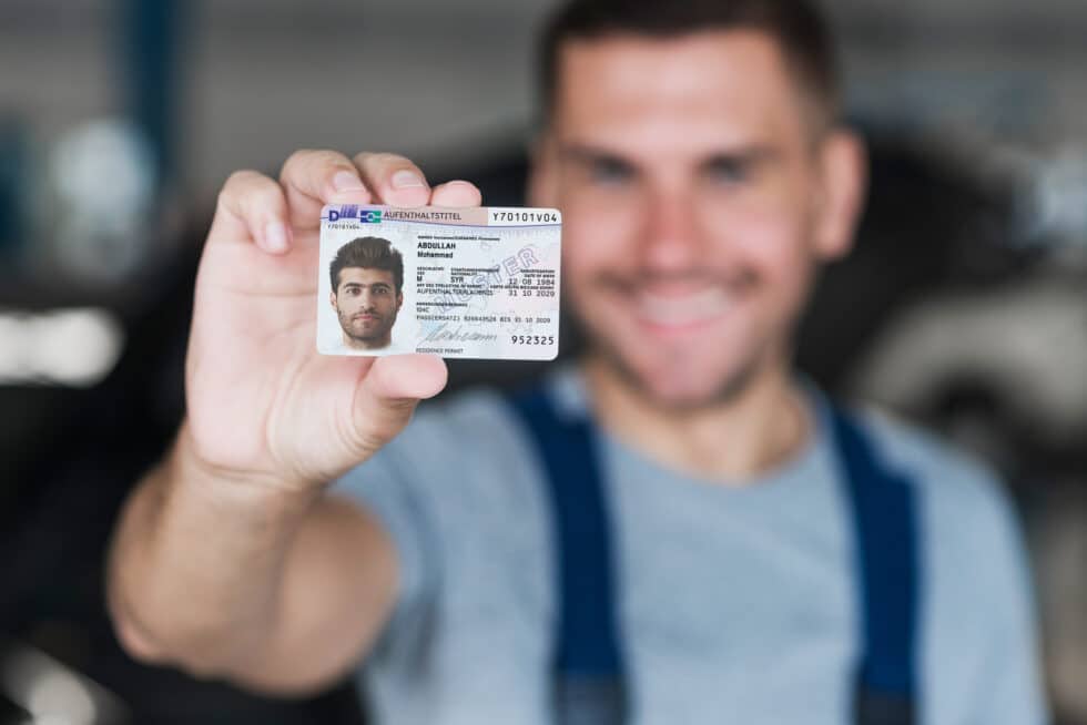 Auf diesem Foto hält ein Mann eine Karte mit dem Aufenthaltstitel 104c AufenthG in der Hand und lächelt.