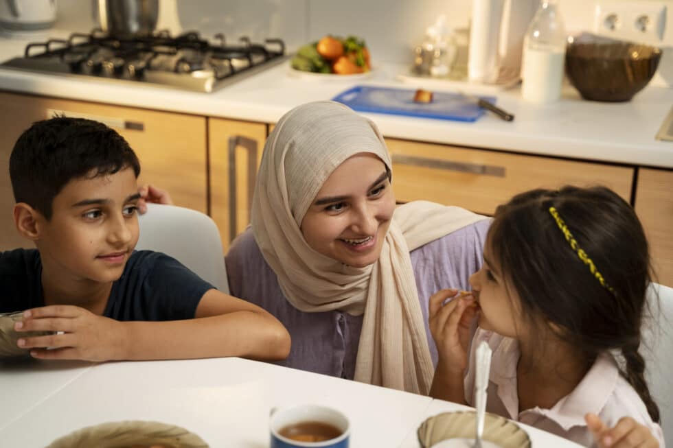 Auf diesem Foto sehen Sie eine muslimische Mutter mit Kindern am Tisch, Im Hintergrund ist eine Küche mit Herd.
