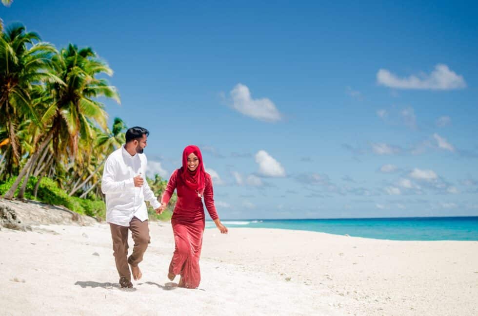 Auf diesem Foto sehen Sie ein muslimisches Paar am Strand. Sie spazieren am Meer und halten sich an der Hand. Der Mann trägt ein weißes Hemd und die Frau ein rotes Kleid und ein rotes Kopftuch. Im Hintergrund sind grüne Palmen
