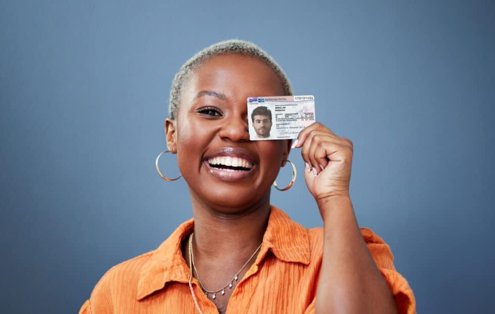 Dieses Foto ist ein Porträt einer Frau die eine Karte mit dem Aufenthaltstitel 25b AufenthG in der Hand hält. Sie verdeckt ein Auge mit der Karte. Die Frau trägt ein oranges Hemd und hat Ohrringe.