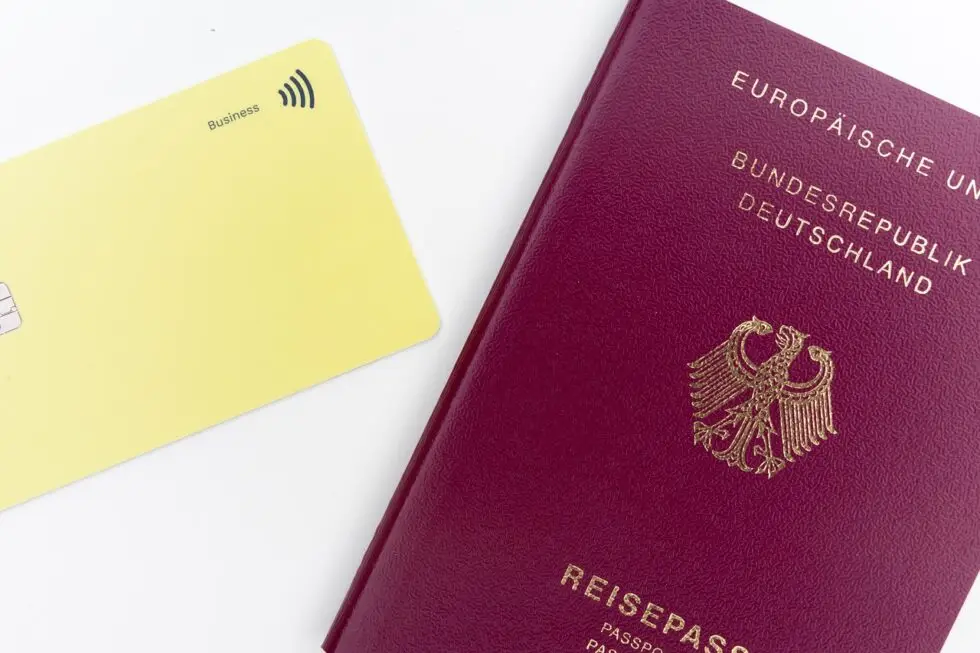 Ein deutscher Reisepass liegt flach auf einer weißen Oberfläche, mit der Vorderseite nach oben gerichtet, sodass das goldene Emblem der Bundesrepublik Deutschland und die Worte "Europäische Union", "Bundesrepublik Deutschland" und "Reisepass" sichtbar sind. Neben dem Pass liegt eine gelbe Business-Kreditkarte mit kontaktlosen Zahlungssymbolen, jedoch ohne sichtbare persönliche Informationen.