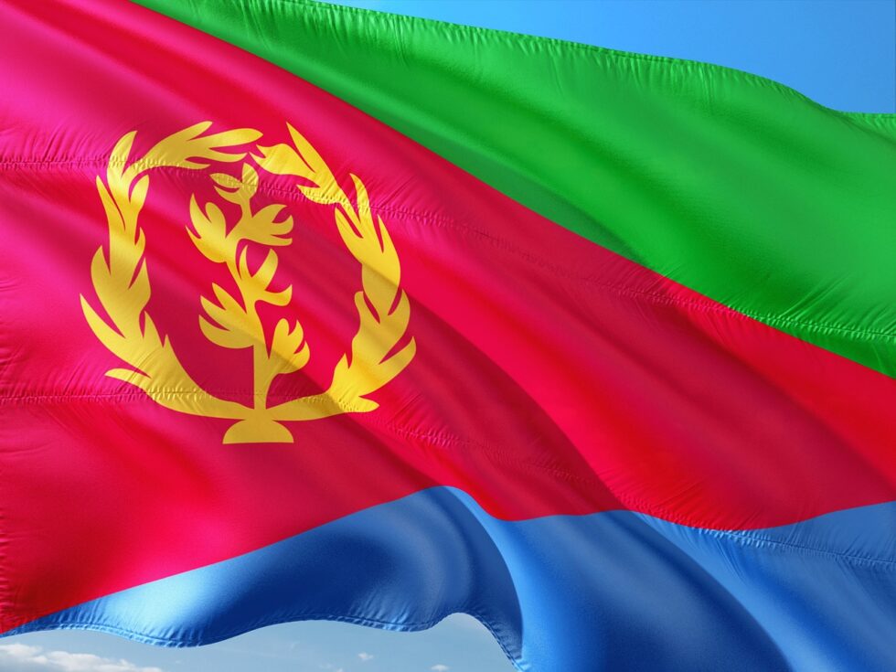 Flag of Eritrea with a green triangle along the mast and red, blue and green horizontal stripes, overlaid by a gold-colored olive branch wreath in the middle.