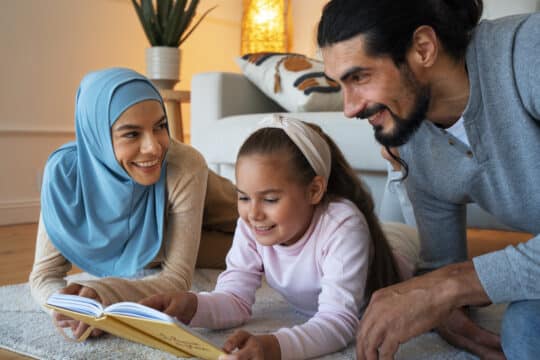 A Muslim couple with their daughter, sitting together on the carpet, smiling and reading a book in a cozy living room.