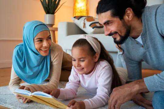 Ein muslimisches Elternpaar mit ihrer Tochter, die zusammen auf dem Teppich sitzen und lächelnd ein Buch lesen, in einem gemütlichen Wohnzimmer.