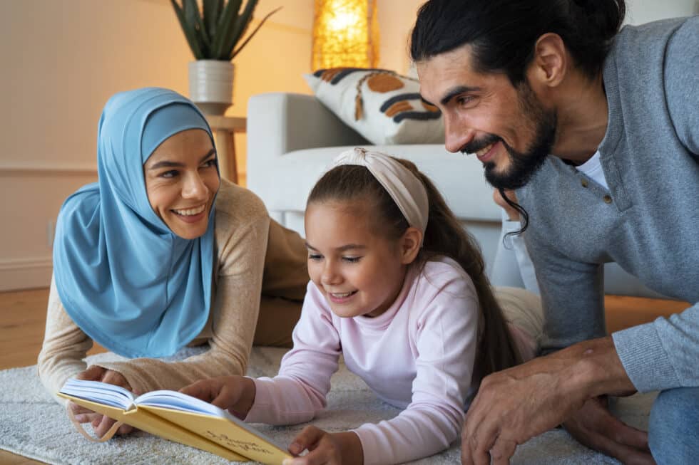 Un couple de parents musulmans et leur fille, assis ensemble sur un tapis et souriant en lisant un livre, dans un salon confortable.