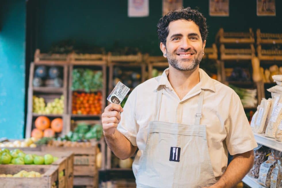 A cheerful vegetable seller in work clothes stands in a grocery store and proudly holds up his German Residence permit , with various types of fruit and vegetables in the background.