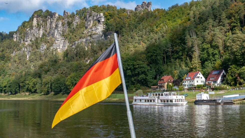 Na pierwszym planie dumnie powiewa niemiecka flaga, a w tle widać tętniącą życiem scenę przedstawiającą rzekę, po której płynie statek pasażerski. Za wodą wznoszą się imponujące, zalesione formacje skalne, a w oddali wzdłuż linii brzegowej widać urocze, kolorowe domy. Obraz oddaje malowniczą naturę i spokojną kulturę mieszkalną Niemiec.