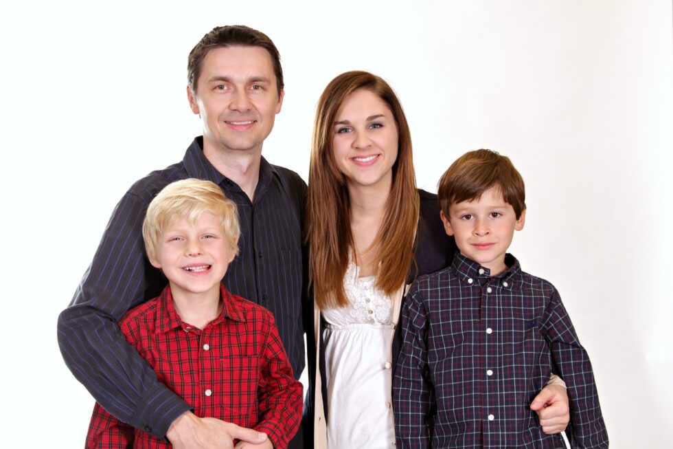 Gülümseyen bir aile portresi; iki yetişkin ve iki çocuk bayramlık kıyafetler giymiş ve mutlu bir şekilde kameraya bakıyor, ikizler kareli gömlek giymiş