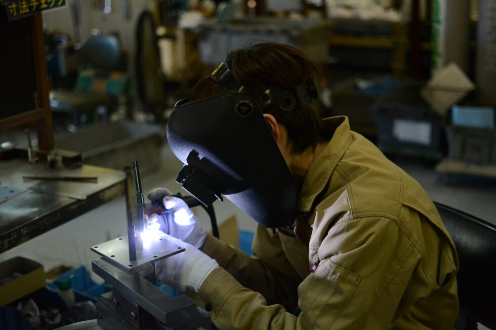 Професионалац у радној одећи обавља посао заваривања машином за заваривање и фокусиран је на рад док носи заштитну маску која прекрива лице и штити очи од интензивне светлости