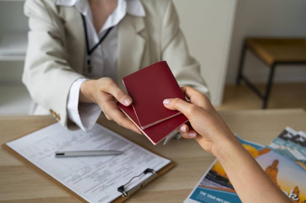 Eine Person im Business-Outfit übergibt einen roten Reisepass an eine andere Person, die am Tisch sitzt, auf dem auch ein Antragsformular und ein Stift liegen, in einer offensichtlichen Büroszenerie.