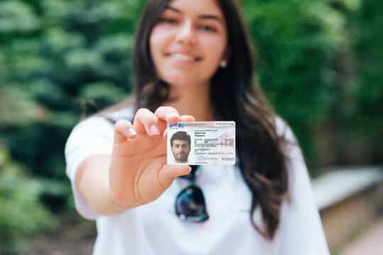 A young woman outdoors proudly holds a German Residence permit § 18c AufenthG into the camera, the focus is on the card against a blurred green background.