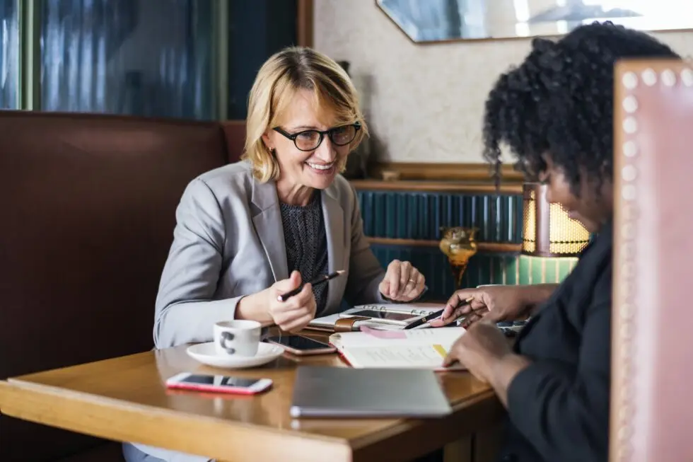 Zwei Geschäftspartnerinnen besprechen Unterlagen in einem Café, möglicherweise beraten sie über den Antragsprozess für einen Aufenthaltstitel in Deutschland.