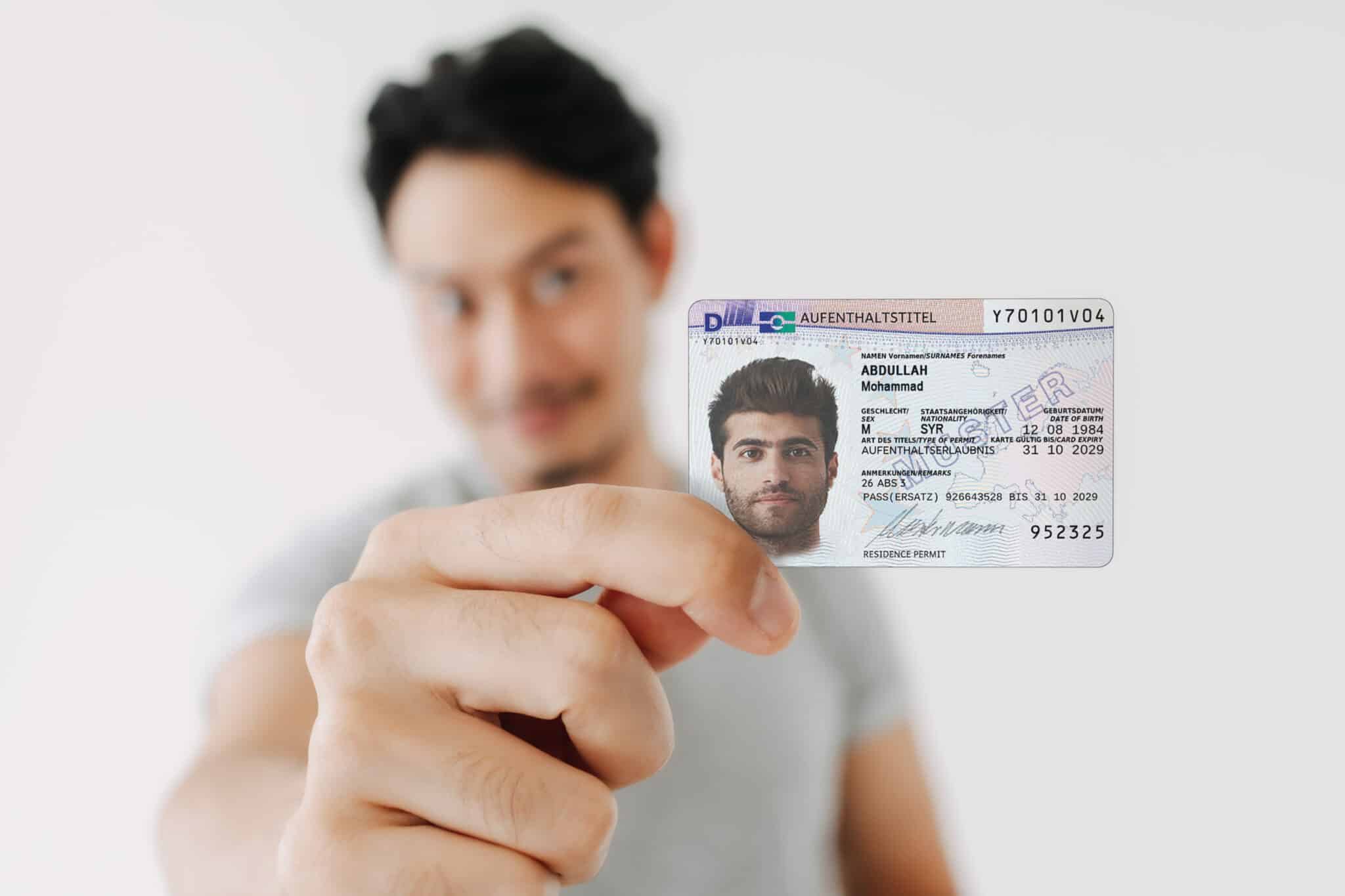 A man smiles and holds a German Residence permit Section 26 (3) AufenthG directly into the camera, with the focus on the card and his face slightly blurred in the background