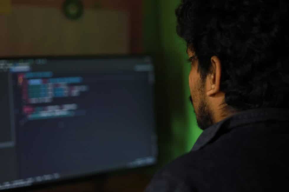 Um especialista em TI, reconhecível pela codificação no ecrã do seu computador portátil, trabalha atentamente numa sala escura com um brilho esverdeado.