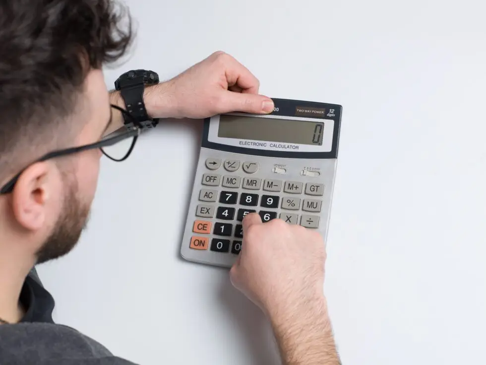 De nombreux demandeurs de naturalisation remarquent au cours du processus que la naturalisation engendre des coûts de toutes sortes : un homme portant des lunettes et une montre noire calcule peut-être ces coûts à l'aide d'une calculatrice de poche argentée. La main droite tape des chiffres tandis que la gauche tient la calculatrice. Seules les mains de l'homme, une partie de la calculatrice et une partie de sa tête sont visibles sur un fond blanc, ce qui met en évidence la concentration du calcul.