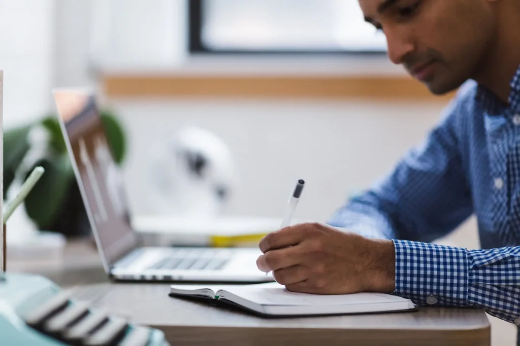 Un homme concentré, vêtu d'une chemise à carreaux bleus, est assis à un poste de travail lumineux et remplit peut-être des documents importants pour la naturalisation. Devant lui se trouve un ordinateur portable ouvert, ce qui montre son engagement dans la recherche et la préparation minutieuse. La scène suggère un environnement calme et organisé, idéal pour remplir des documents officiels.