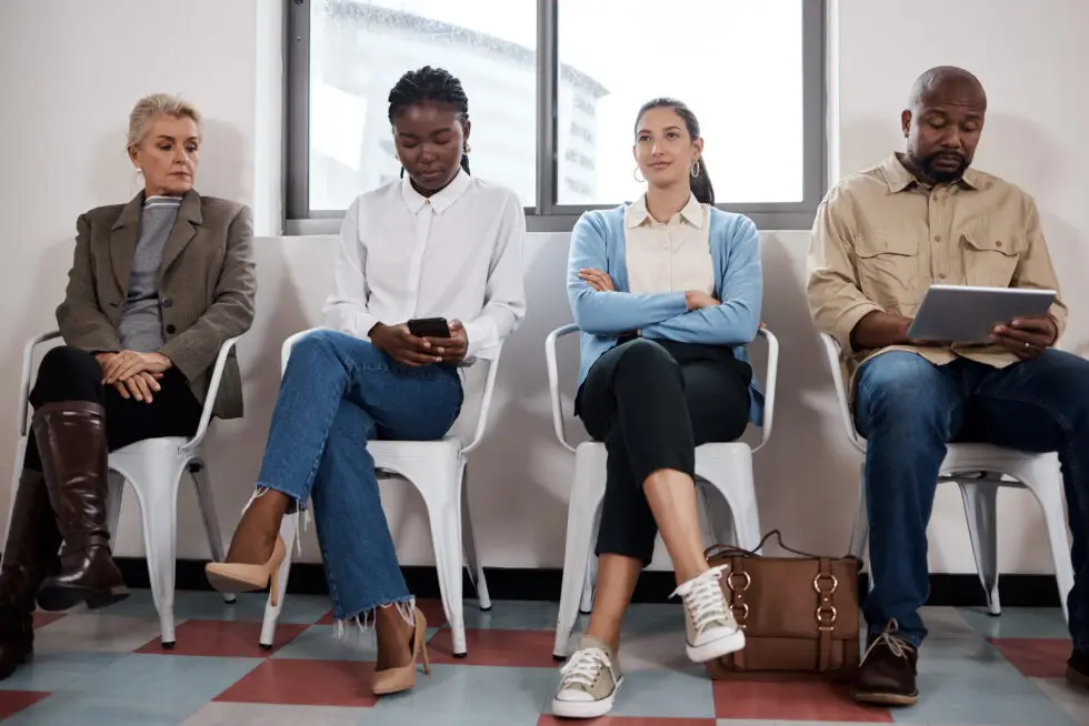 Einbürgerung und Termin: Eine Gruppe von Menschen wartet in einem modernen Büro auf ihre Gesprächstermine zur Einbürgerung. Eine ältere Dame und drei weitere Personen, jeweils in professioneller Kleidung, sitzen nebeneinander. Sie schauen in verschiedene Richtungen, einige beschäftigt mit digitalen Geräten, was auf eine ruhige, geduldige Atmosphäre hindeutet.