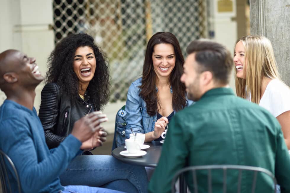 A cheerful, multicultural group of five friends are enjoying a coffee break together. They are sitting outside at a small round table, surrounded by a lively city atmosphere. The joy and laughter they share convey a sense of community and friendship.