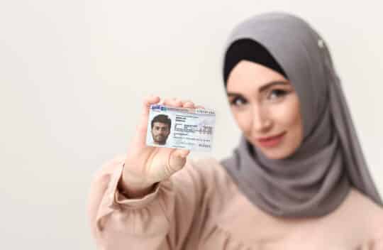 一名头戴头巾的妇女微笑着拿着德国居留证§ 23 AufenthG 对着镜头，焦点在卡片上，背景中她的脸被模糊了。
