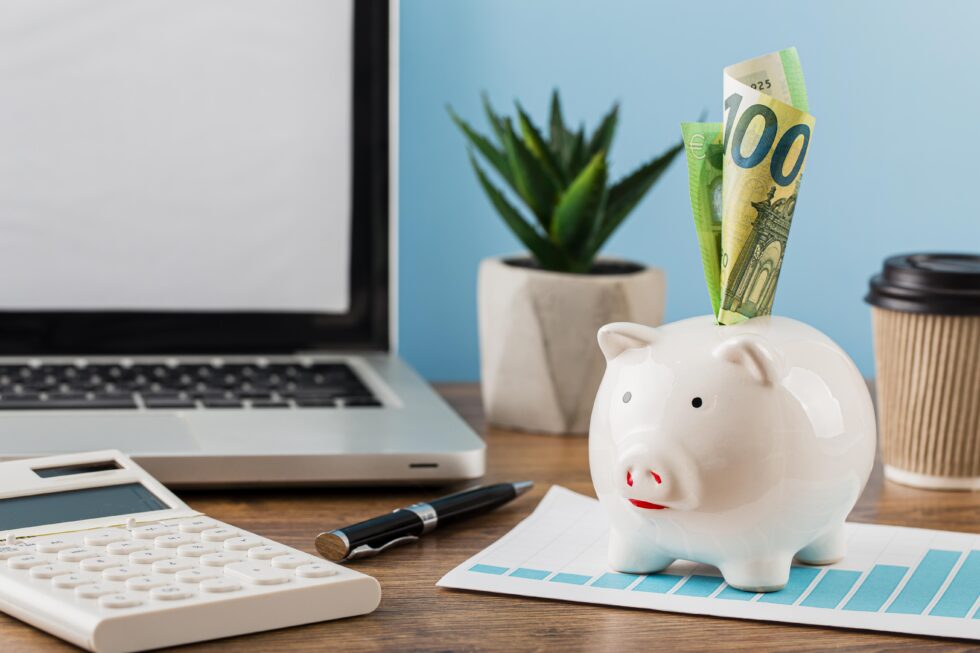 A workplace with a focus on financial planning and savings, represented by a transparent piggy bank on a desk with a 100 euro bill sticking out of it. Next to it is a laptop with a white screen, a calculator, a coffee mug, a pen, a green potted plant in the background and a growth chart on paper in front of the piggy bank, which could indicate financial growth or budget planning.