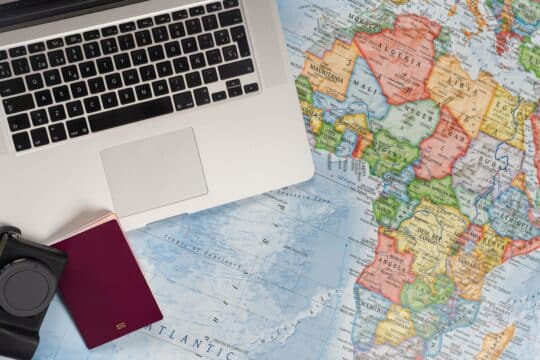 Un spațiu de lucru pentru planificarea naturalizării, cu un laptop, un aparat foto și un pașaport roșu pe o hartă detaliată a lumii care evidențiază continentul african. Această configurație simbolizează pregătirea pentru călătorii internaționale sau studierea proceselor de naturalizare din diferite țări.