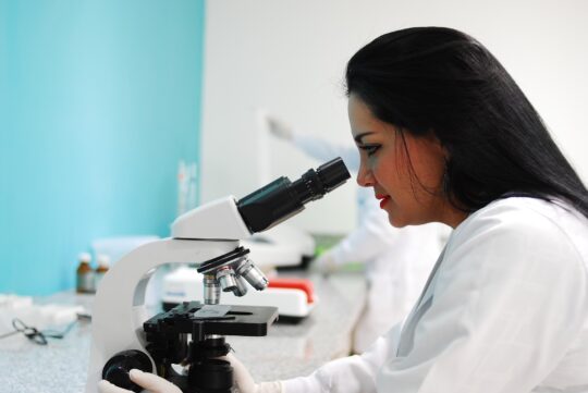 A scientist, possibly an EU Blue Card holder, in a white lab coat is focused on examining samples through a microscope. The clear and bright laboratory environment with turquoise walls emphasizes the clean and precise working environment. Her commitment to research reflects the expertise promoted by the EU Blue Card in Europe.