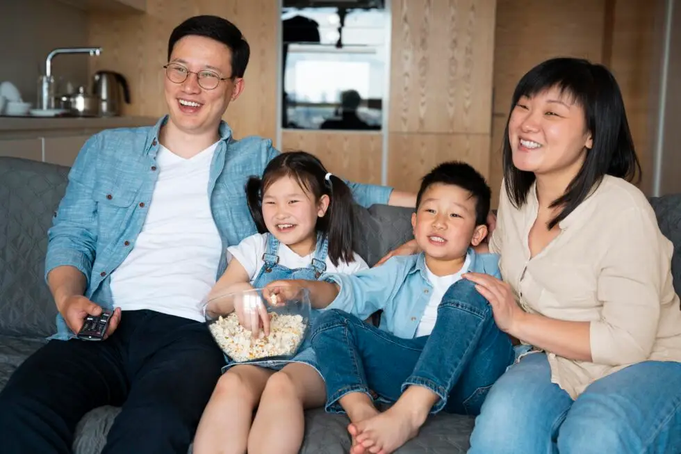 Eine glückliche asiatische Familie sitzt zusammen auf einer Couch in einem modernen Wohnzimmer. Der Vater, der eine Brille und ein blaues Jeanshemd trägt, sitzt neben seiner Tochter, einem kleinen Mädchen im Jeanskleid, das lächelnd in eine Schale Popcorn greift. Neben ihnen sitzt der Sohn in einem passenden blauen Hemd, und die Mutter, die ein beiges Oberteil trägt, umarmt den Jungen liebevoll. Alle lachen und genießen offensichtlich eine gemeinsame Zeit.