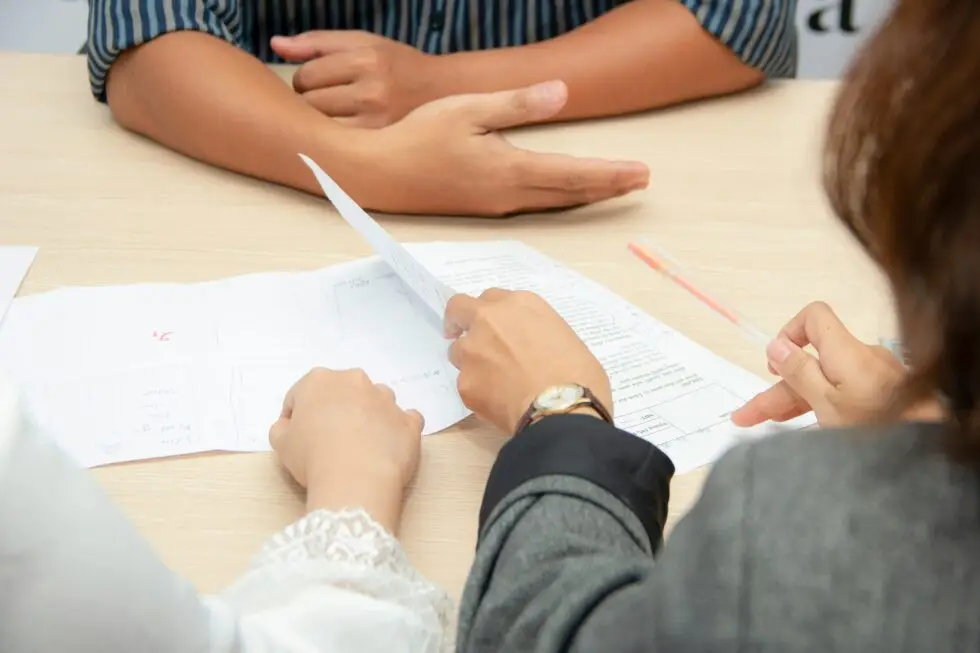 Drei Personen sitzen an einem Tisch und konzentrieren sich auf das Ausfüllen und Überprüfen von Dokumenten, wobei ihre Hände und die Papiere im Vordergrund stehen, während ein Stift und weitere Unterlagen auf dem Tisch liegen