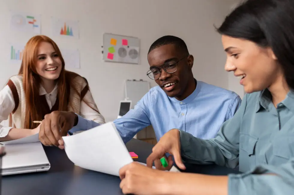 Drei junge Berufstätige, eine rothaarige Frau und zwei Personen mittlerer Hautfarbe, sitzen an einem Tisch und diskutieren lächelnd über ein Dokument, was Teamarbeit und soziale Integration am Arbeitsplatz symbolisiert.