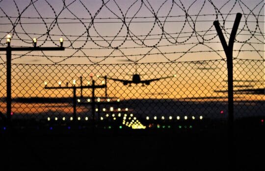 日落时分，一架飞机在铁丝网后起飞。这一画面象征着根据《居留法》第 54 条，因某些刑事犯罪或对公共安全的威胁而引发的驱逐意向，驱逐外国人。